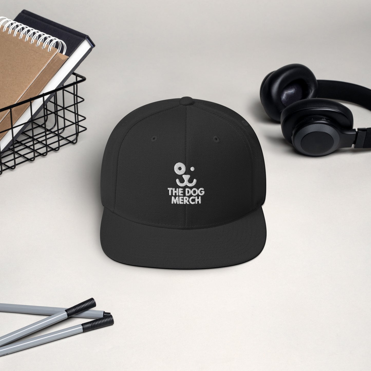 Snapback hat that says The Dog Merch color Black front side, headphones, notebooks and pens in the background 