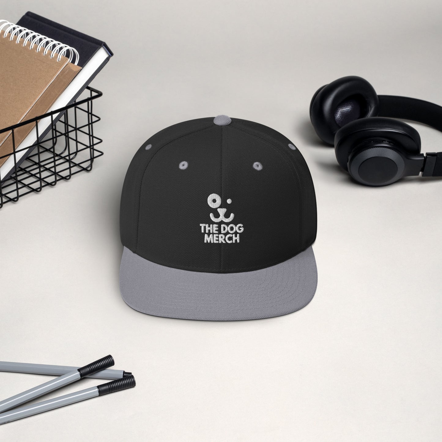 Snapback hat that says The Dog Merch color Black Silver front side, headphones, notebooks and pens in the background 